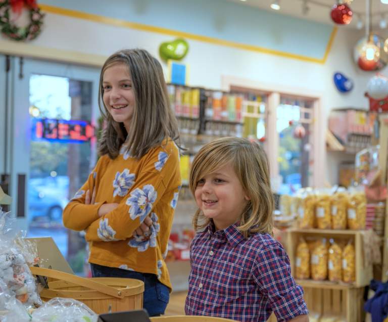 Two smiling children stand at the counter of a colorful candy store, surrounded by jars of sweets and holiday decorations.