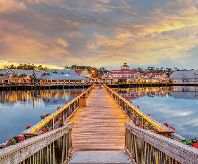A wooden boardwalk decorated with holiday garland stretches over calm water toward Barefoot Landing in North Myrtle Beach at sunset, with shops and lights reflecting on the surface.