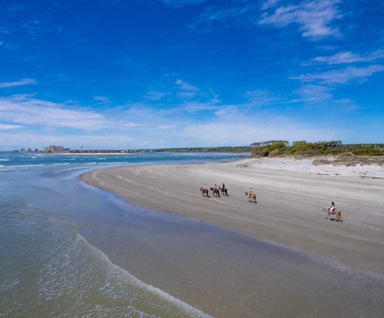 A group horseback rides on the beach of Waites Island with North Myrtle Beach in the distance
