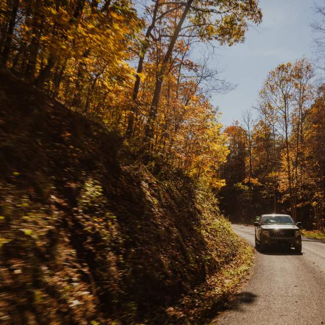A truck drives along a paved road during a sunny day with fall foliage along the roadside.