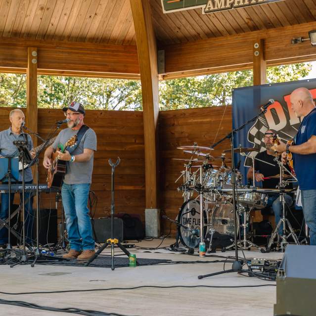 Three members of a band play instruments on an outdoor covered stage.