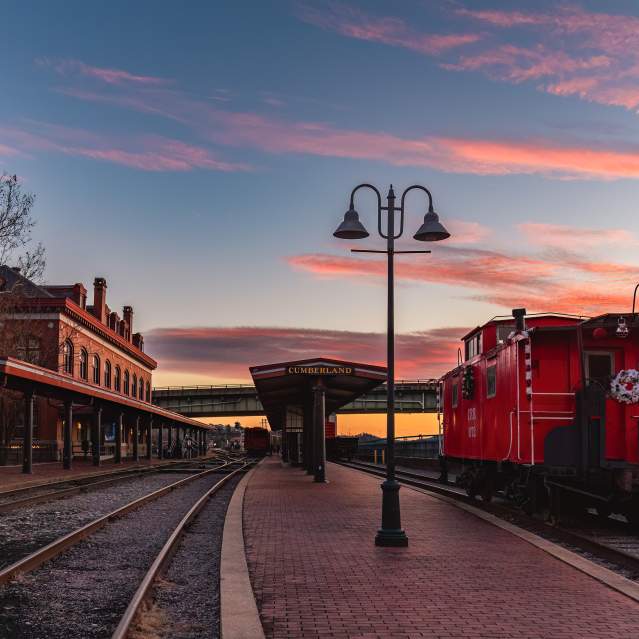 A red train caboose with a holiday wreath sits parked on train tracks in front of a historic train station at sunset.