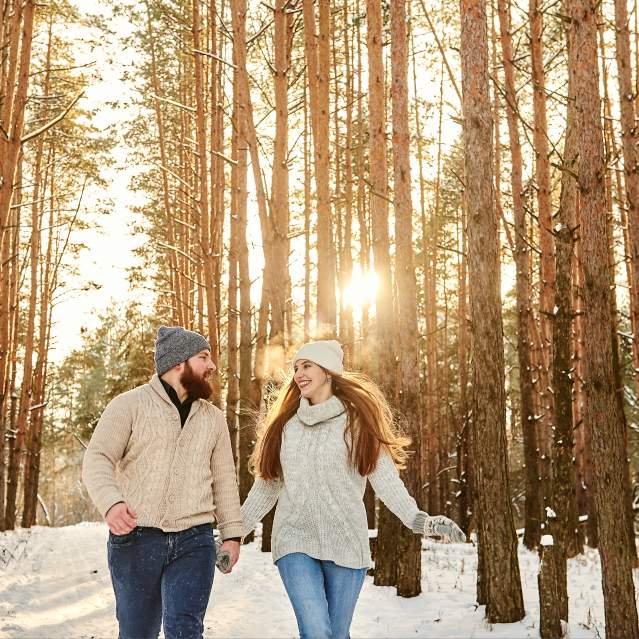 A man and woman hold hands and walk through the snow covered forest.