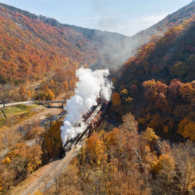 A steam train travels through a mountain valley surrounded by vibrant autumn foliage.