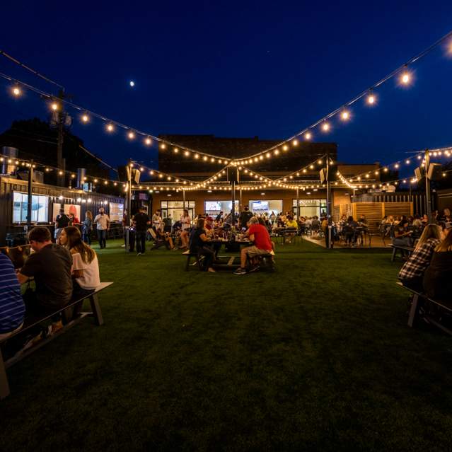 Image is of Covington Yard at night with twinkle lights above, people sitting at table and food trucks (operating out of containers) is lining the area.