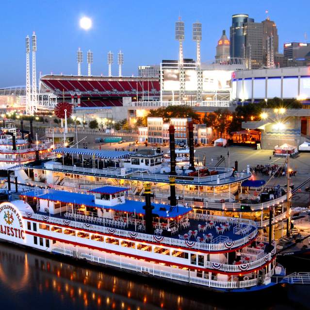 Several sternwheeler riverboats on the Cincinnati shoreline in 2006. It's evening, the boats and buildings are lit up as the sky grows darker.