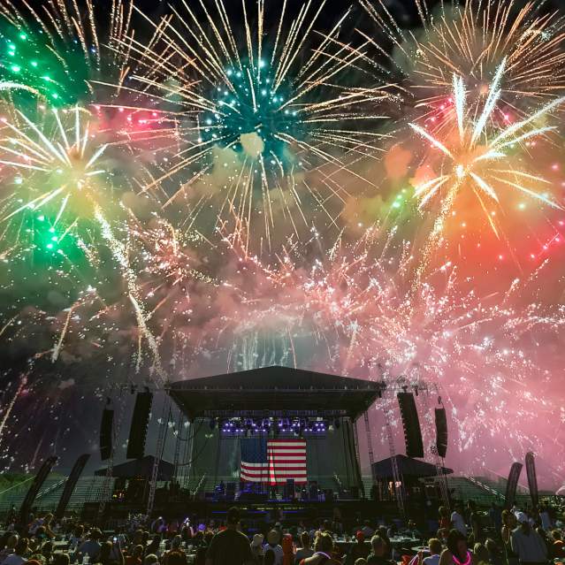 A colorful Four of July fireworks display in the sky above a stage that has the American flag hanging as the backdrop