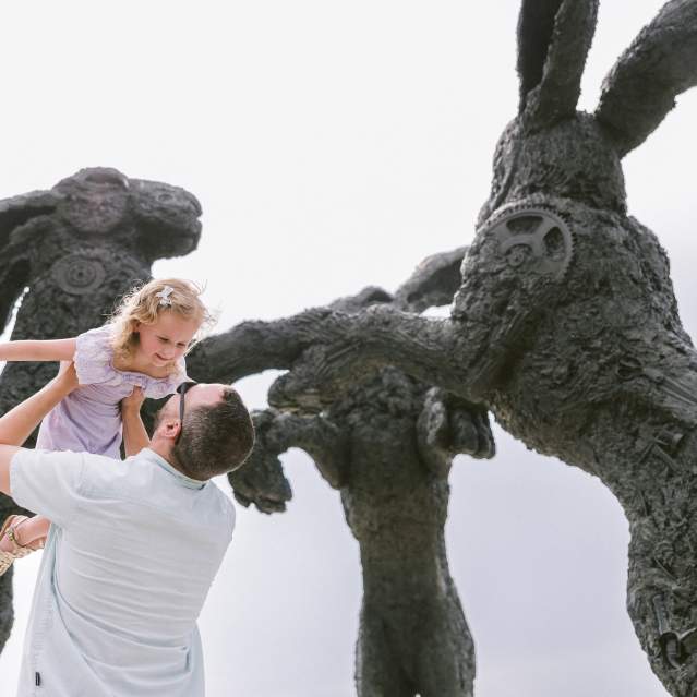 A father lifting his smiling daughter in front of large, textured rabbit sculptures in a whimsical outdoor setting, under a bright sky.