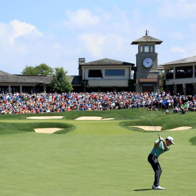 Golfer swining club at golf ball towards the 18th green that is surrounded by a crowd at the Memorial Touranemnt.