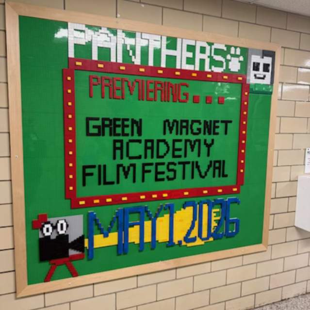 A bulletin board made of green LEGO baseplates mounted on a tan brick wall. The board features pixelated text and graphics constructed from LEGO bricks. At the top, the word "PANTHERS" is spelled out in white bricks next to a white paw print. The central text, framed by a red and yellow border, reads "PREMIERING... GREEN MAGNET ACADEMY FILM FESTIVAL." In the bottom left, a LEGO movie camera icon appears to project a yellow beam of light toward the date MAY 1, 2026.