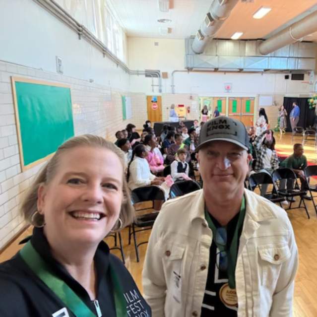 A selfie of a Kim Bumpas and Curt Willis standing in a school gymnasium. Curt is wearing a grey baseball cap with "FILM KNOX" printed on it and a white denim jacket. In the background, a large audience of students is seated in black folding chairs, and a stage with green curtains is visible in the distance. The room features high ceilings with industrial ductwork and tan brick walls.