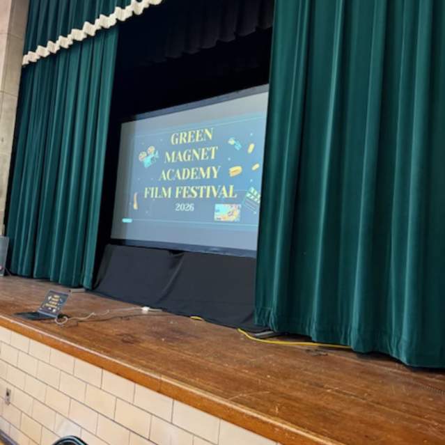 A view of a school stage framed by heavy green curtains. A large projection screen in the center displays a title slide for the "GREEN MAGNET ACADEMY FILM FESTIVAL 2026." The slide features yellow text on a dark background accented with small icons of movie cameras and film strips. A laptop and several power cables are visible on the wooden floor at the base of the stage.