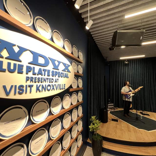 A man in a white shirt plays guitar on a stage with a blue curtain backdrop, surrounded by a blue wall with a "WYDX Blue Plate Special" sign and a "Visit Knoxville" banner.