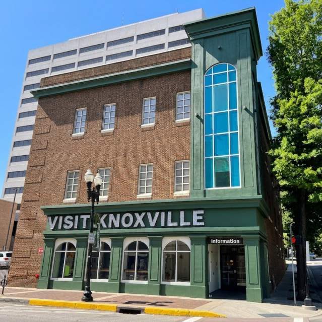 A bright, daylight photo of the Visit Knoxville building on a city street corner under a clear blue sky. The three-story building features a red-brick exterior and a prominent green-painted corner section with a large, vertical arched window. The words "VISIT KNOXVILLE" are displayed in large, white marquee-style letters across the green ground-floor facade, which also has several large arched windows. A black vintage-style street lamp stands on the sidewalk in front. In the background, a tall, white multi-story building rises to the left, and a lush green tree borders the street to the right. The entire building facade is uniformly lit with no visible shadows.