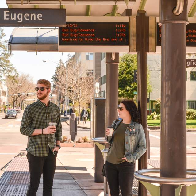 A man and a woman holding coffee in paper cups stand at the bus stop under the sign "Eugene".