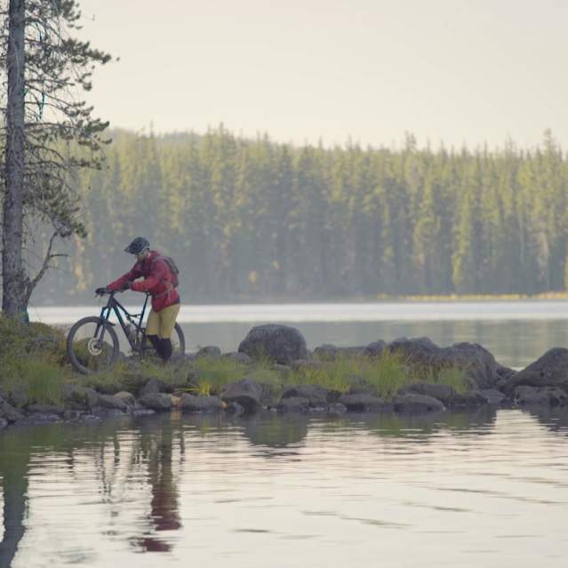 A person walks a bike along the rocky shoreline of a misty lake.