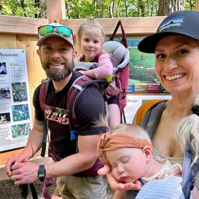 Ready for a spring hike, a young couple stands in front of informational signs at Hawk Mountain Sanctuary. The man wears a backpack holding his young daughter and the mother wears a harness that holds a sleeping little girl.