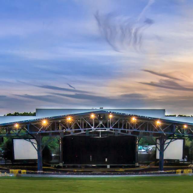 an outdoor concert venue, there is a large tent with seating underneath it as well as a lawn. There are trees in the background behind the tent/stage.