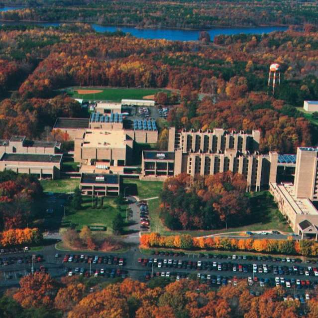 a birds eye view of a campus with buildings, a parking lot filled with cars, surrounded by trees with fall foliage
