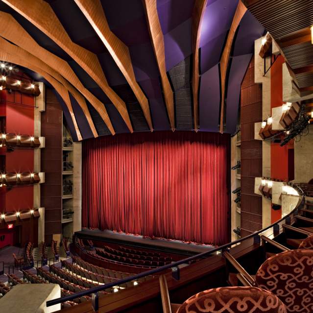 inside a theater, the picture is taken from the side of "stage left" on the balcony, looking towards the stage which has a red curtain