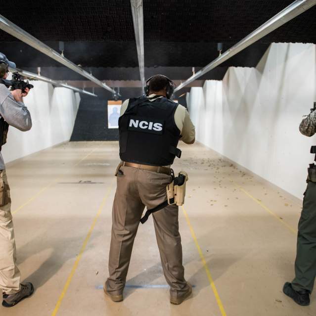 3 men pointing away from the camera, all with black vests on. The 2 men on the left have vests that say "NCIS" and the man on the right has "federal agent" written on the back of his vest. They are holding guns at an indoor gun range.