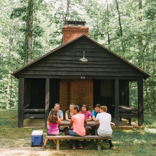 7 people sitting at a picnic bench in front of a cabin in a forest