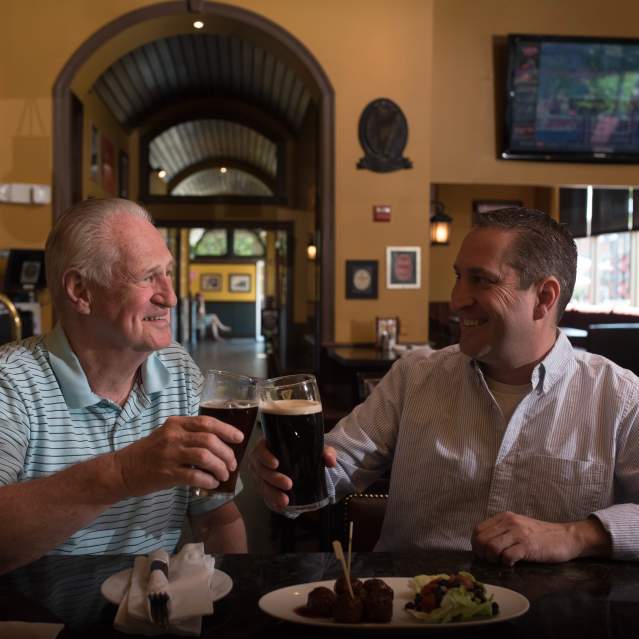 2 men smiling, drinking beer, sitting at a bar with a plate of food on the bar