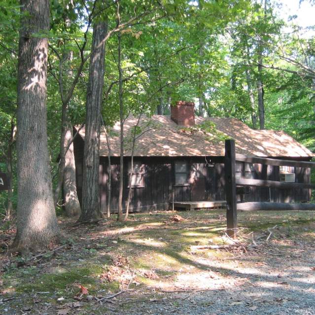 a brown cabin in a forest, surrounded by trees, there is a sign in front of the cabin that says "orenda"