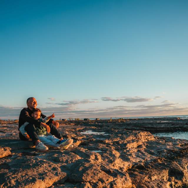 An adult and child sit together on a rocky shoreline at sunset, looking out over calm water. Warm golden light illuminates the scene as the child points toward the horizon beneath a wide blue sky streaked with soft clouds.