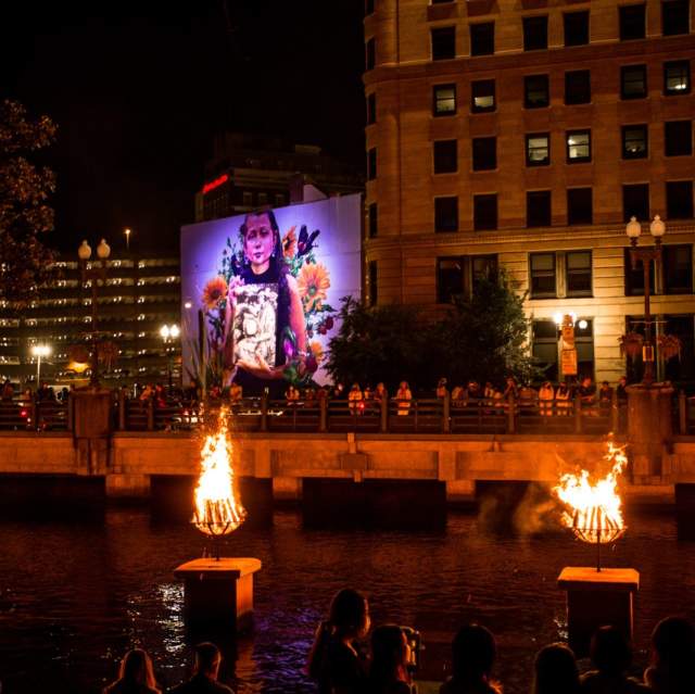 Braziers burn on the river and a mural of a young girl is illuminated in the background.