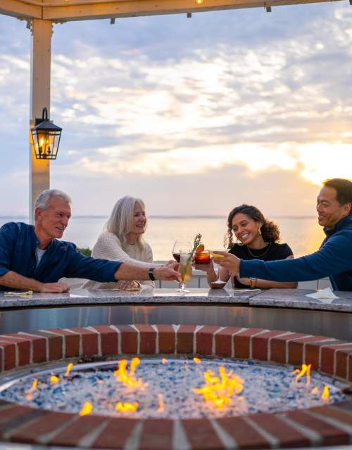 Four people gather around a circular fire pit, toasting drinks as the sun sets over a serene waterfront. Lanterns illuminate the scene.