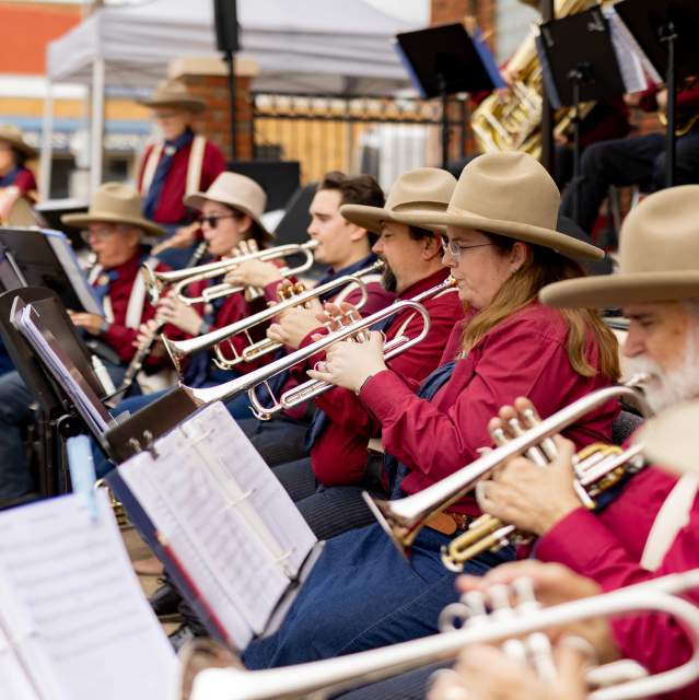 The trumpet section of the Buffalo Bill's Cowboy Band play their instruments at the 2026 Doc Holliday Festival. They are wearing red long sleeve button up shirts and brown felt cowboy hats. Sheet music is displayed on the music stands in front of the musicians.