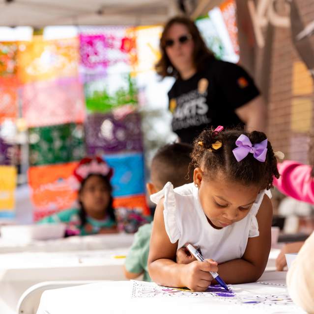 Young children coloring at an outdoor community event with colorful papel picado banners in the background.
