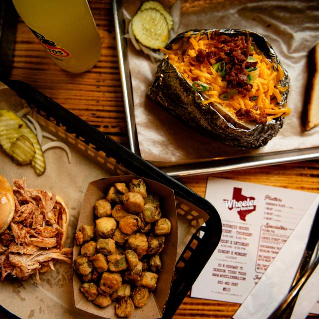 Two trays of food from Wheeler Barbeque viewed from above. One tray has a pulled pork sandwich with the top bun set to the side, a side of fried okra, and a couple of sliced pickles on the side. The other tray shows a baked potato loaded with cheese, green onions, and bacon with a slice of bread on the side.