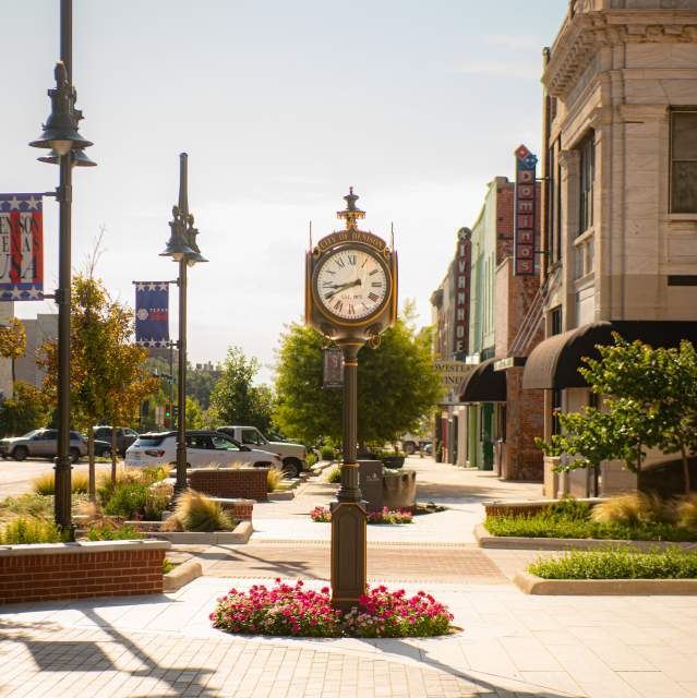 The City of Denison clock that is located in front of City Hall. It is a bright sunny day with clear blue sky. Pink flowers are in the bed around the base of the clock.