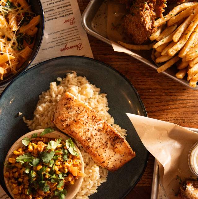 A top down view of a table filled with food at the George Tavern. The central plate has a piece of baked salmon on a bed of rice with a side bowl of Mexican street style corn. The bowl in the upper left corner is filled with a pasta dish. The upper right tray contains some chicken tenders and a pile of French fries.