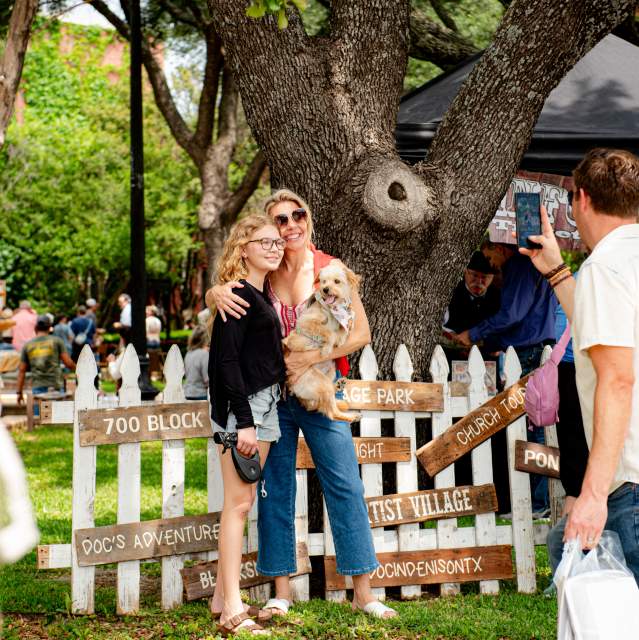 A blonde middle aged woman stands with one arm holding a small dog and the other arm draped over the solder of a younger girl with blonde curly hair. They are smiling and standing in front of a white picket fence decoration at the Doc Holliday Festival and a man in a white shirt is taking a picture of them with a cell phone.