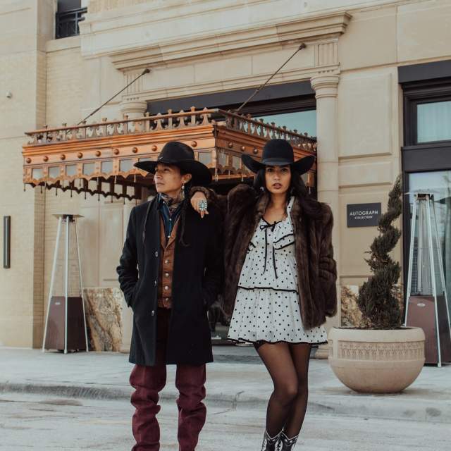 A stylish duo stands outside an elegant historic building, embodying Western Black Tie fashion. The man wears a black cowboy hat, a tailored black overcoat over a brown vest and scarf, deep red trousers, and polished boots, accessorized with turquoise jewelry. The woman exudes modern Western glam in a polka-dot mini dress with a fur coat, sheer tights, black-and-white cowboy boots, and a black hat. Their confident stance, set against the backdrop of an ornate hotel entrance, captures Amarillo’s blend of high fashion and rugged Western heritage.