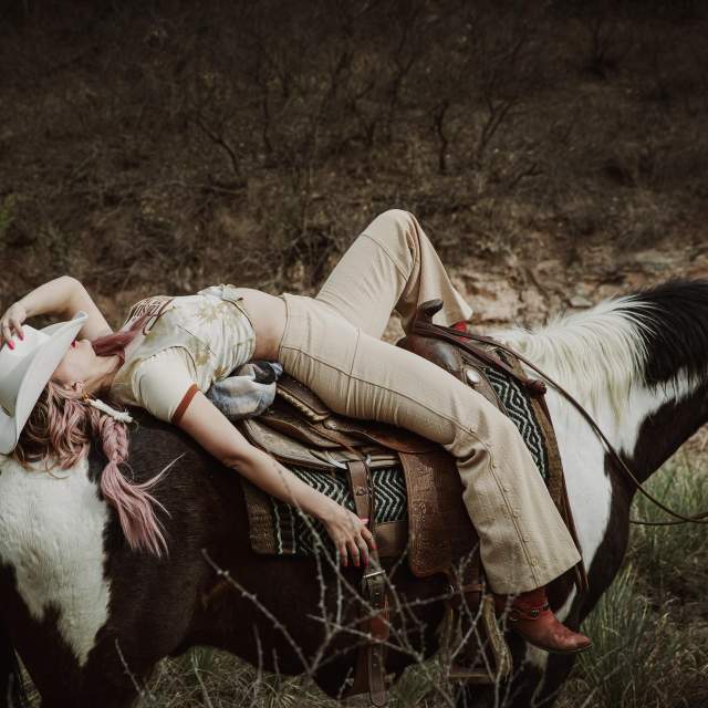 ChatGPT said:

A woman with pink braids and a white cowboy hat reclines effortlessly on a black-and-white horse, her hand shading her face as she rests against the saddle. She wears beige bell-bottoms and red boots, surrounded by rugged brush and canyon terrain that capture Amarillo’s untamed Western spirit.