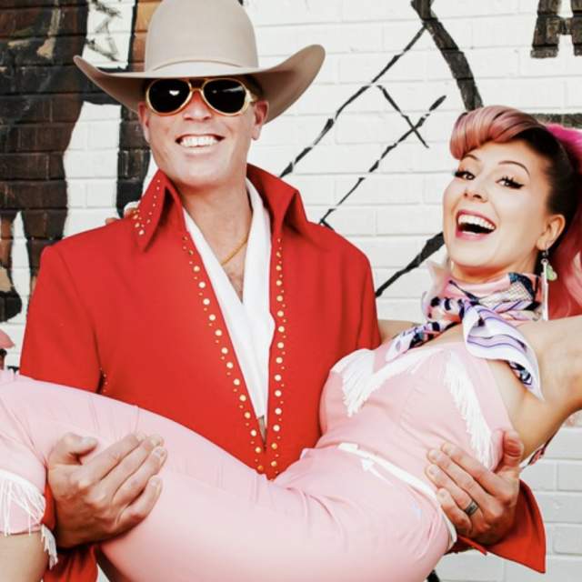 Two lively performers dressed in vibrant retro outfits pose in front of a mural celebrating Amarillo's Route 66. The mural features iconic imagery, including a classic red truck and the Route 66 shield.