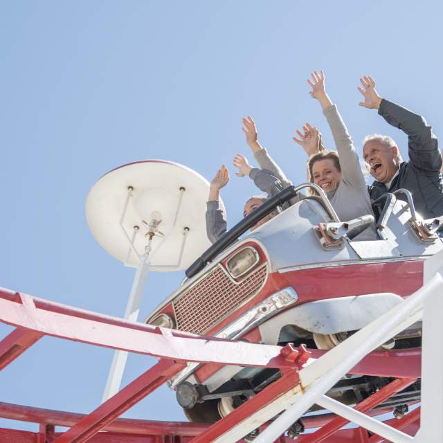 thrill-seekers of all ages throw their hands in the air as a red-and-white roller coaster car dives along its track under a bright blue Amarillo sky
