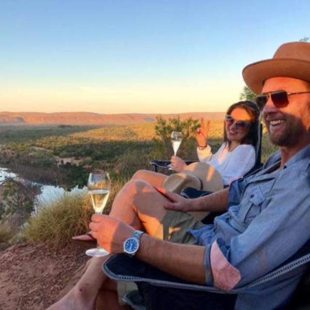Two people sitting at a scenic lookout point at sunset, holding glasses of sparkling wine