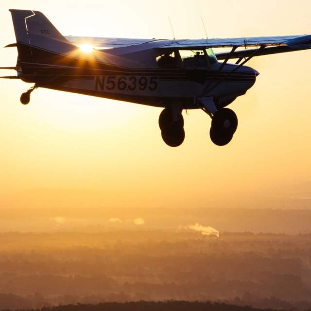 Silhouette of a small airplane, registration N56395, flying at sunrise with golden light illuminating the hazy landscape below.