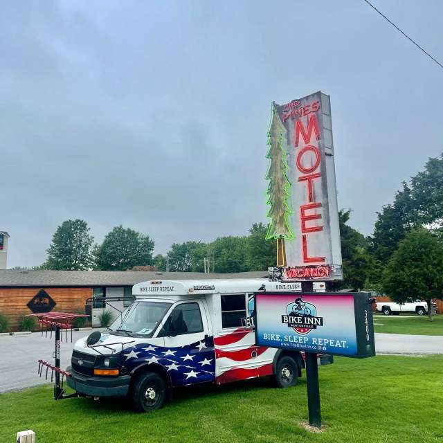 American flag-themed shuttle bus in front of 'BIKE INN' sign with a neon-lit 'PINES MOTEL' sign overhead on an overcast day.