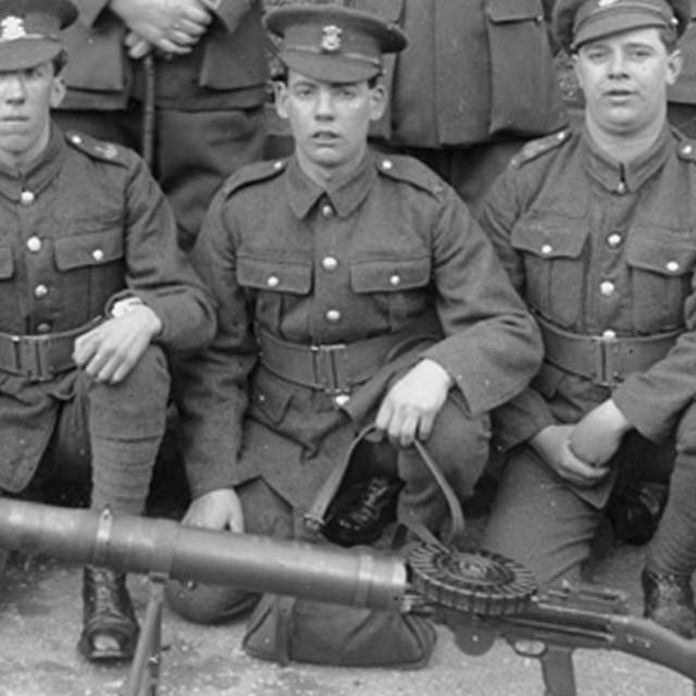 A Welsh regiment of soldiers kneel behind a Lewis Gun. A row of soldiers stand behind them with only their feet visible. The soldiers are in First World War army uniforms and hats.