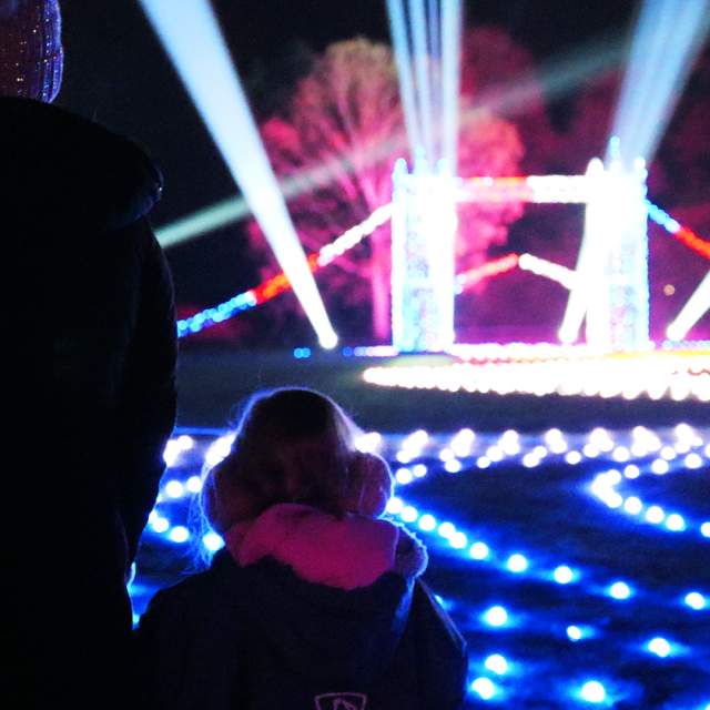 A mother and small child stand with their backs to the camera looking at a small, lit up London Bridge. There are roaming spotlights & glowing fairylights on the floor in all different colours. The trees off in the distance are also lit up in blue and pink.