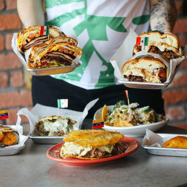 A person offering the viewer tortas in both hands with a table of tortas, empanadas and more food below