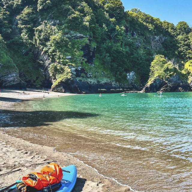 A serene cove with turquoise waters, surrounded by lush green cliffs. People are kayaking and relaxing on the sandy shore under a clear blue sky.