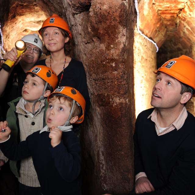 A family in hard hats explores a dimly lit tunnel, looking amazed as an adult shines a flashlight. The scene conveys curiosity and adventure.