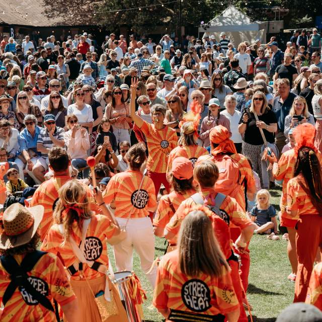 A lively crowd watches performers in bright orange costumes playing instruments at an outdoor festival. The atmosphere is energetic and festive.